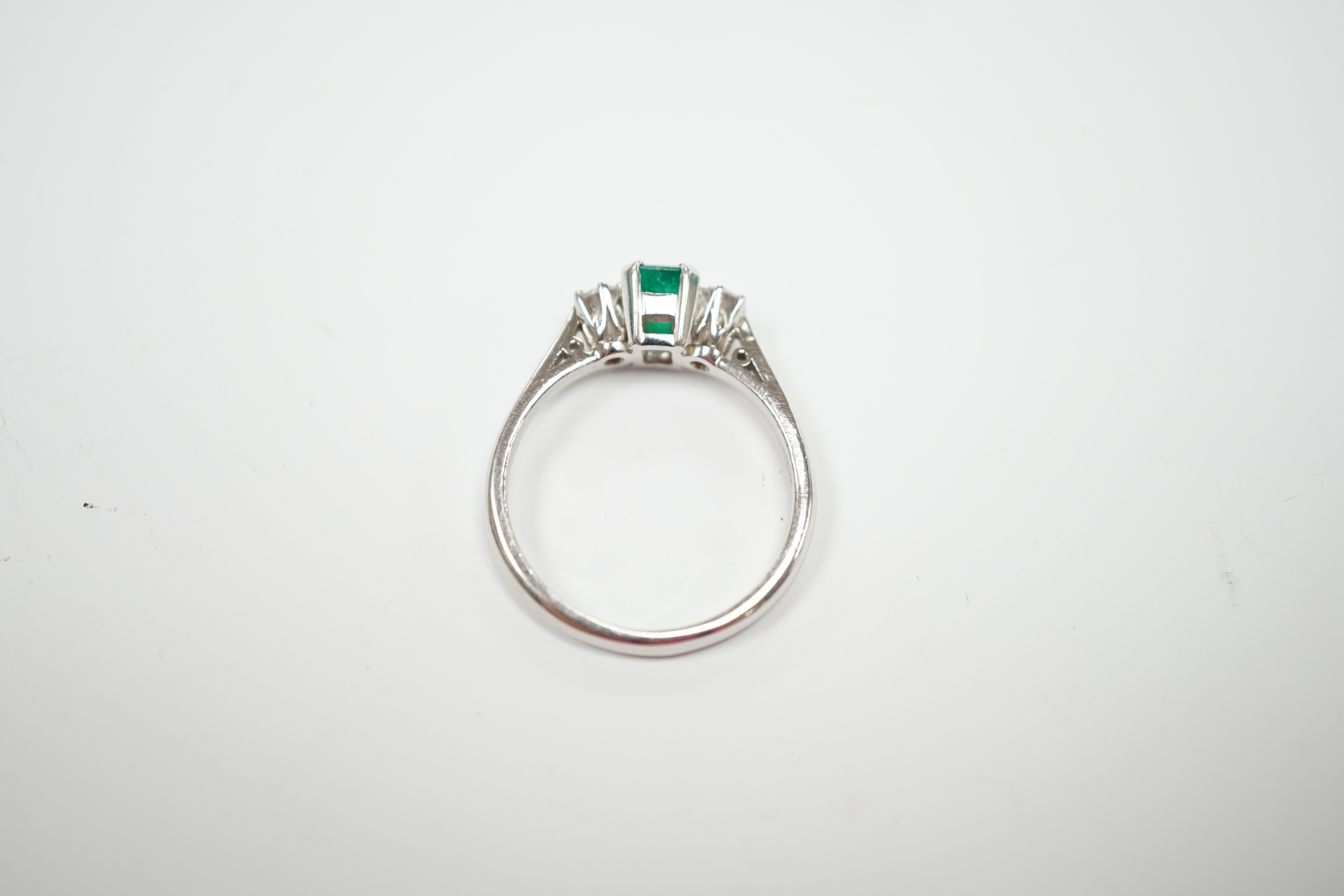 A modern 18ct white metal, single stone emerald and two stone diamond set ring, size N, gross weight 4 gram.